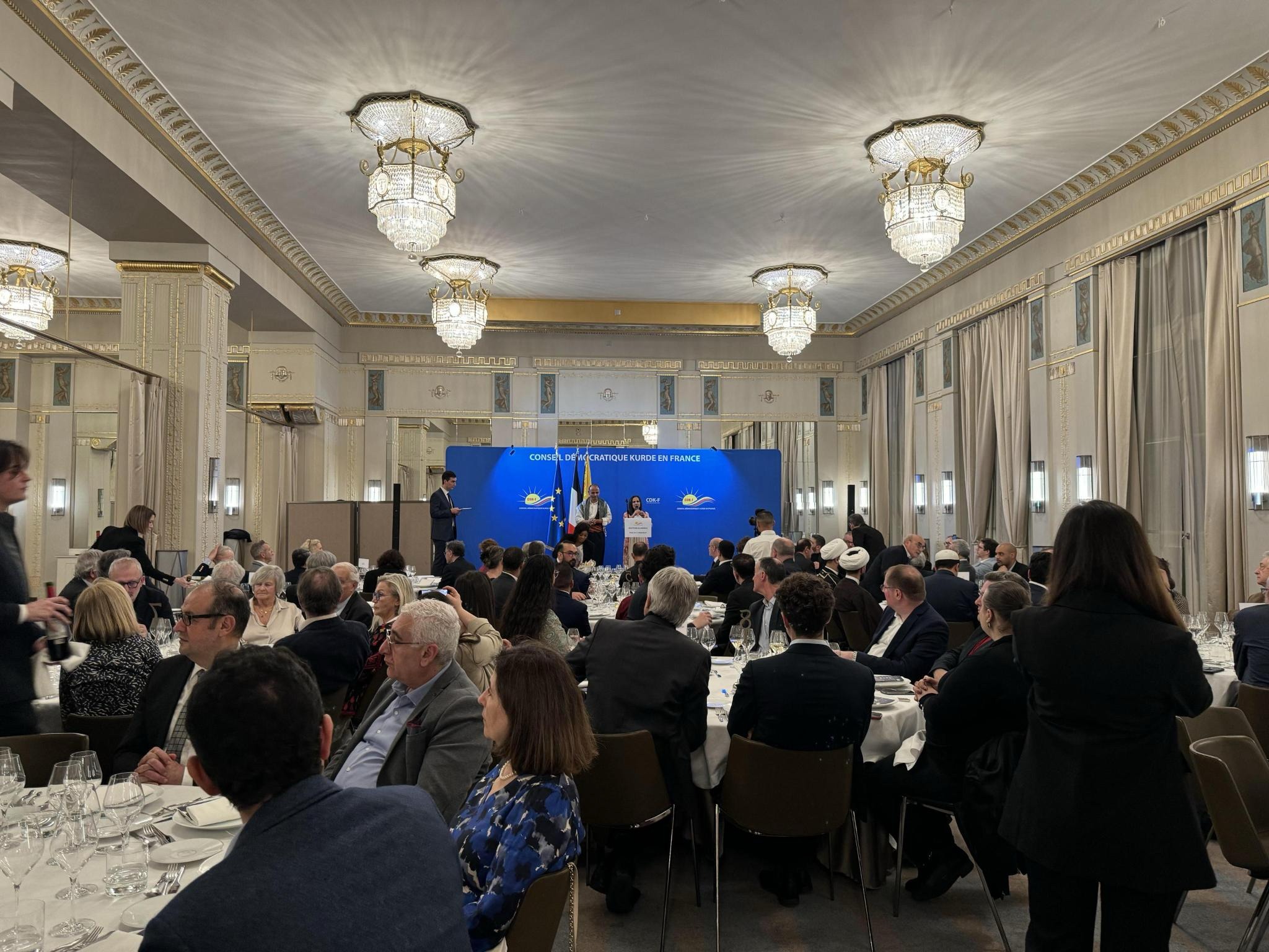 Rencontre du CDK-F dans une salle de réception, avec une prise de parole devant les drapeaux et le panneau institutionnel